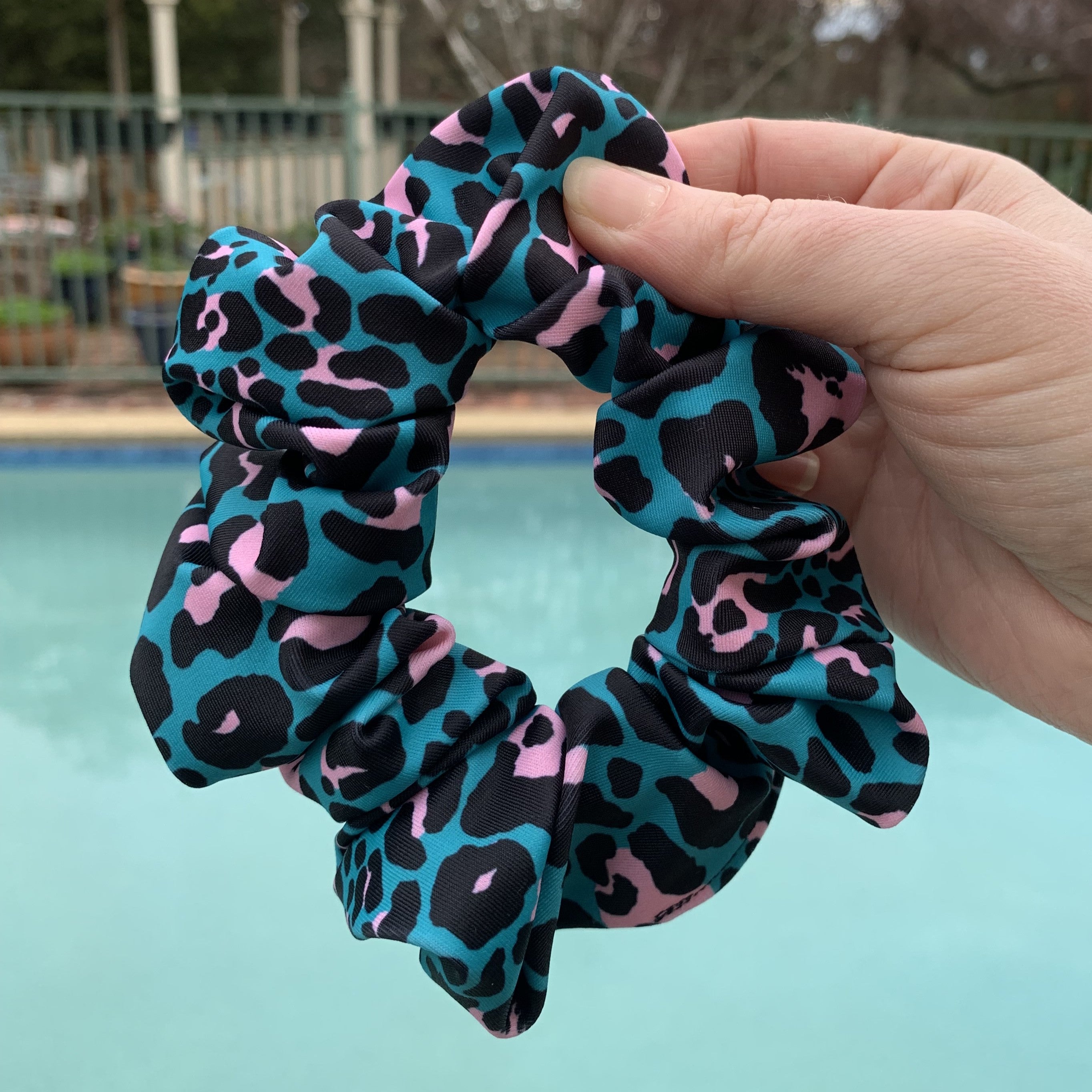 Hand holding a blue and pink leopard print swim scrunchie with a pool in the background