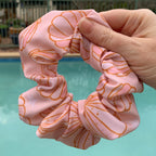 Pink swim scrunchie with shell pattern held by a hand against a poolside background