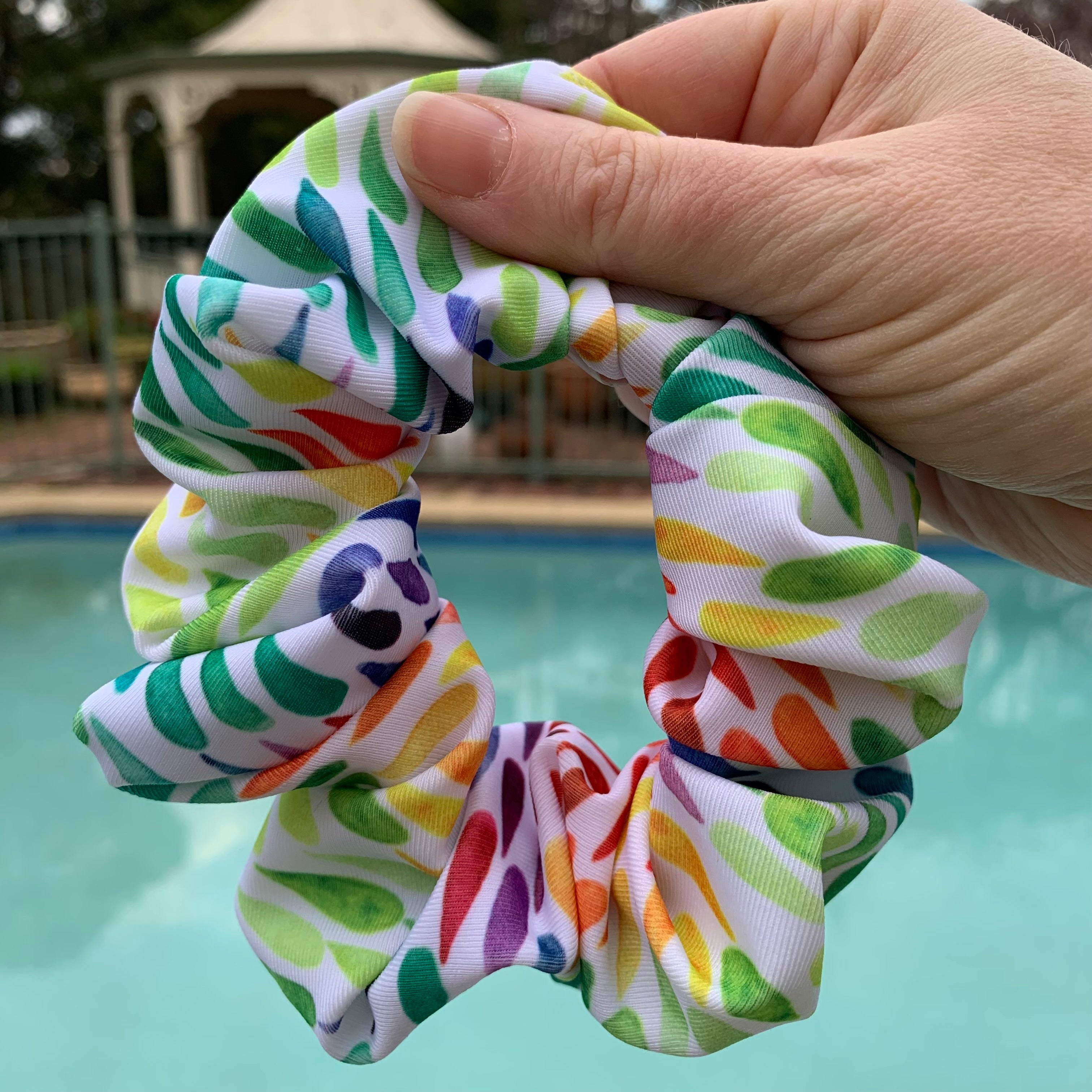 Colourful patterned swim scrunchie held by a hand with a pool in the background