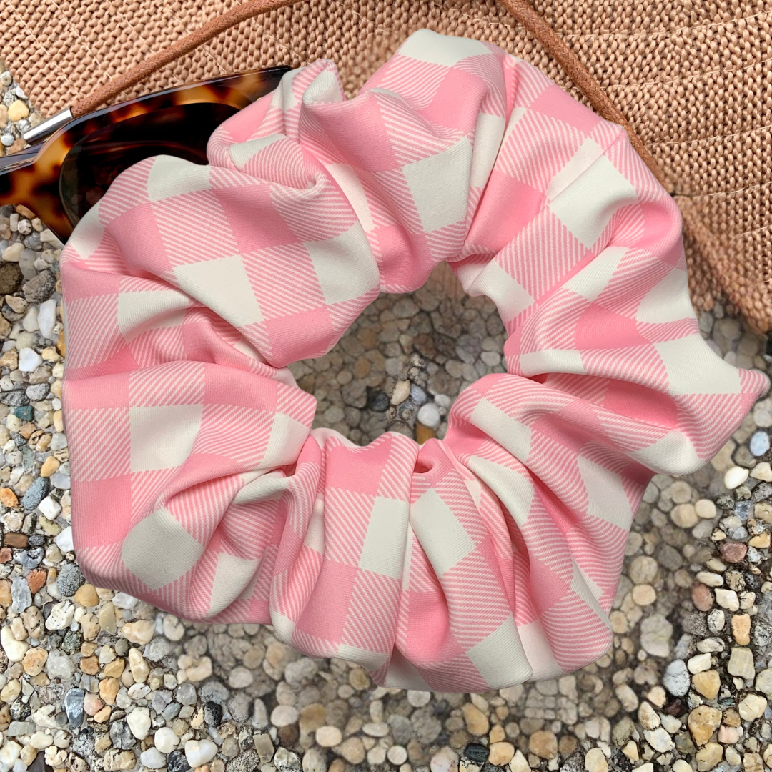 Pink and white checkered swim scrunchie on a pebbly surface with a hat and sunglasses