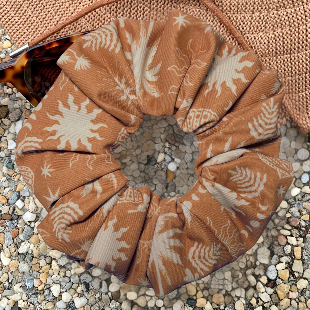 A fabric scrunchie with a brown and cream botanical sunburst pattern, styled on a bed of small pebbles with a woven straw hat and sunglasses in the background.