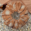 A fabric scrunchie with a brown and cream botanical sunburst pattern, styled on a bed of small pebbles with a woven straw hat and sunglasses in the background.