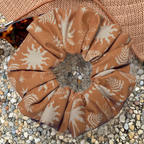 A fabric scrunchie with a brown and cream botanical sunburst pattern, styled on a bed of small pebbles with a woven straw hat and sunglasses in the background.