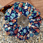 A fabric hair scrunchie with a colorful floral print on a navy blue background, styled on a bed of small pebbles next to a hat and sunglasses.