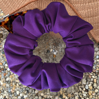 A purple fabric scrunchie, displayed on a bed of small pebbles with a brown woven hat and a partial tortoiseshell eyeglass visible in the corner.