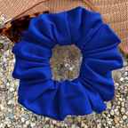 A bright royal blue fabric scrunchie laid flat on a pebble surface, with part of a brown woven hat and tortoiseshell sunglasses visible around it.