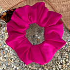 A bright hot pink fabric scrunchie, displayed on a bed of small beige pebbles with a woven straw hat and sunglasses in the background.