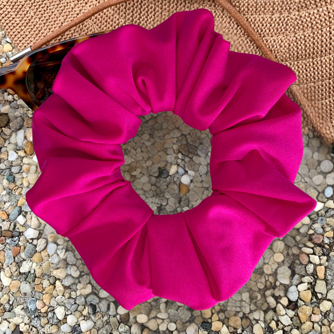 A bright hot pink fabric scrunchie, displayed on a bed of small beige pebbles with a woven straw hat and sunglasses in the background.