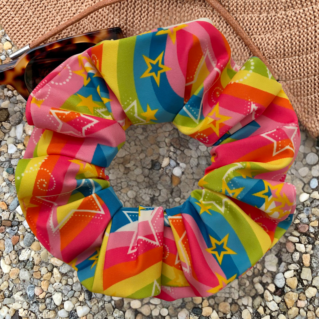 A colorful fabric hair scrunchie with a rainbow stripe pattern and yellow star motifs, styled on a pebble surface next to sunglasses and a woven bag.