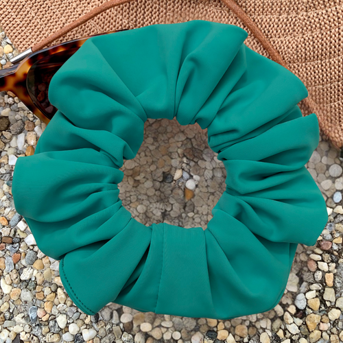 A scrunchie in a vibrant teal green color, styled on a bed of small pebbles with a woven straw hat and tortoiseshell sunglasses partially visible in the background.