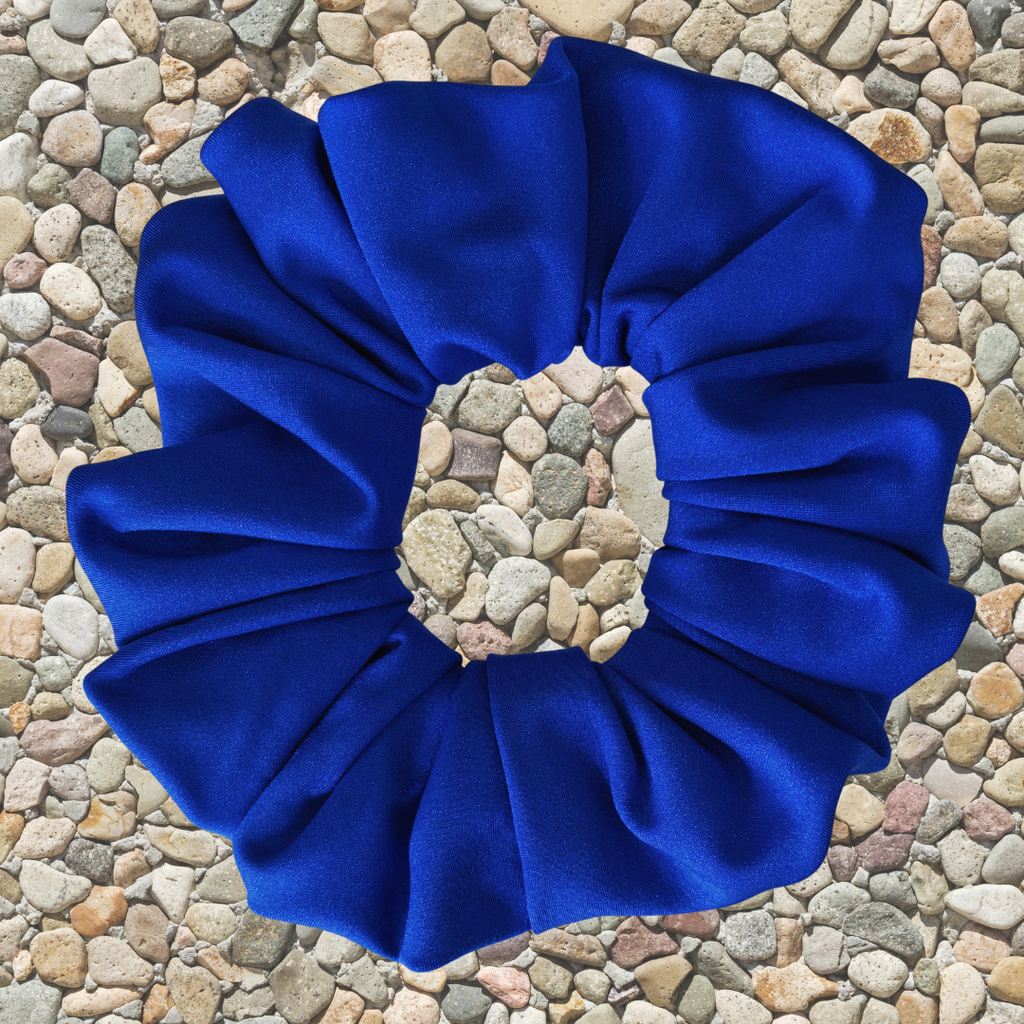 A royal blue fabric scrunchie with a gathered, pleated texture, laid flat on a background of small multicolored pebbles.