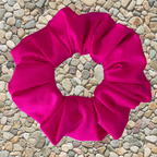 A bright hot pink fabric scrunchie laid flat on a background of small beige pebbles.