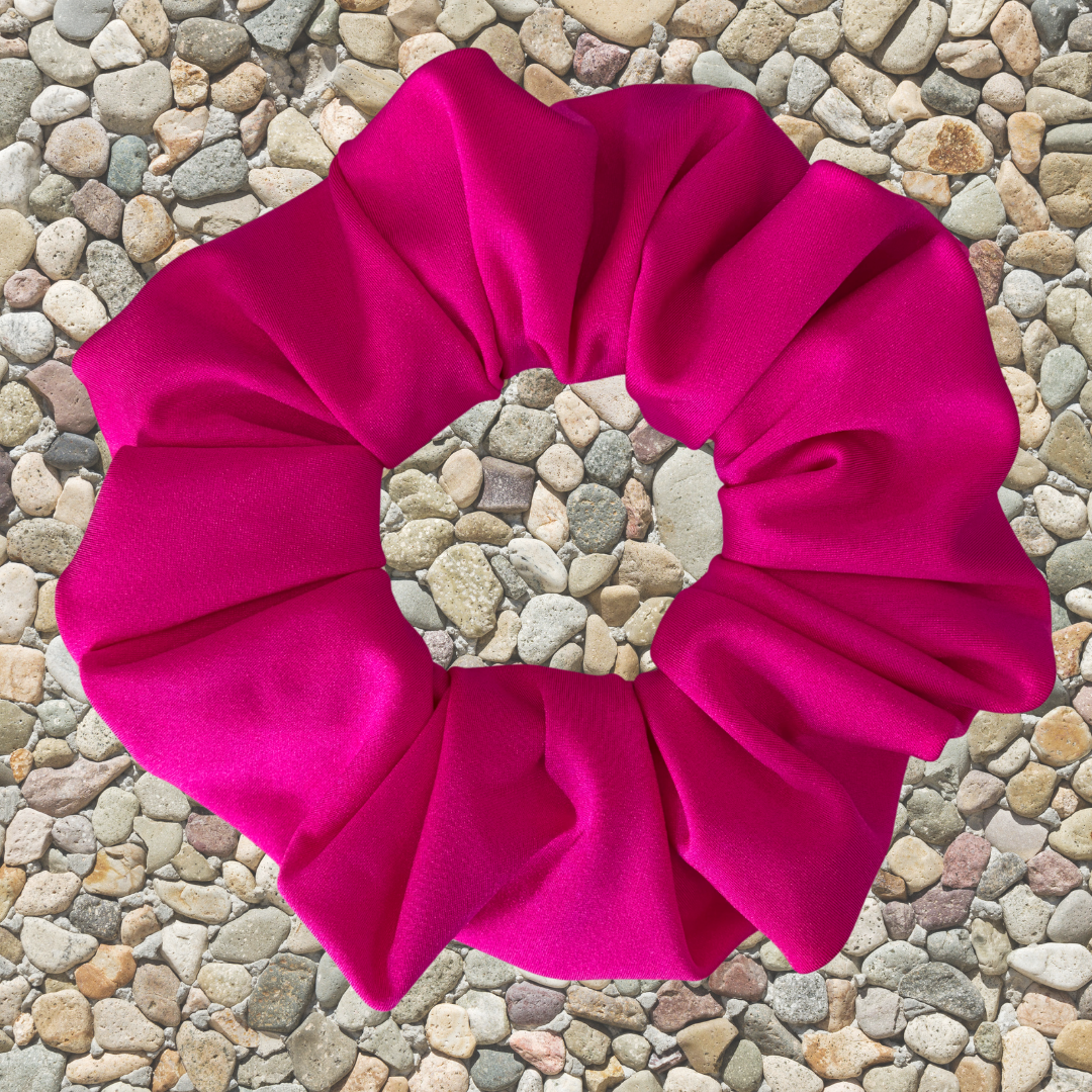 A bright hot pink fabric scrunchie laid flat on a background of small beige pebbles.