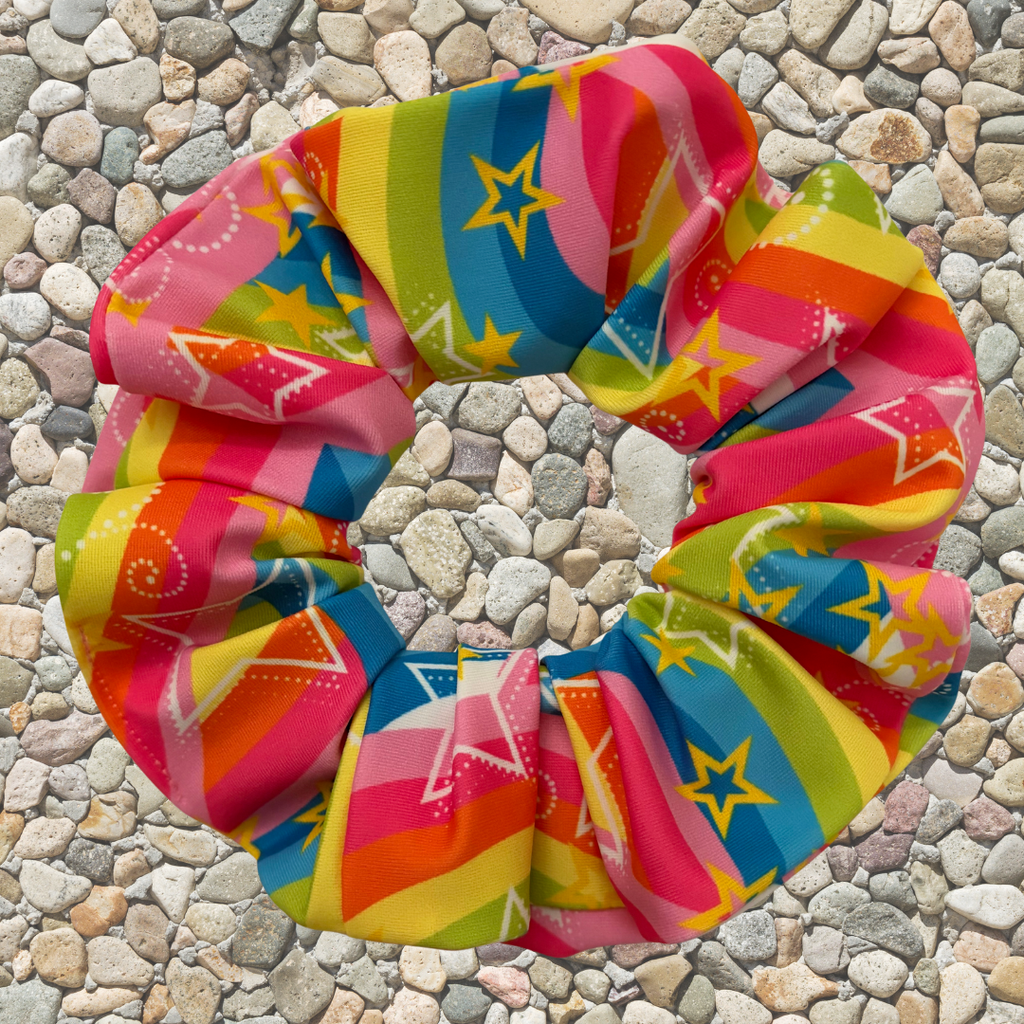 A colorful fabric hair scrunchie with a rainbow stripe and star pattern, laid flat on a background of small gray pebbles.