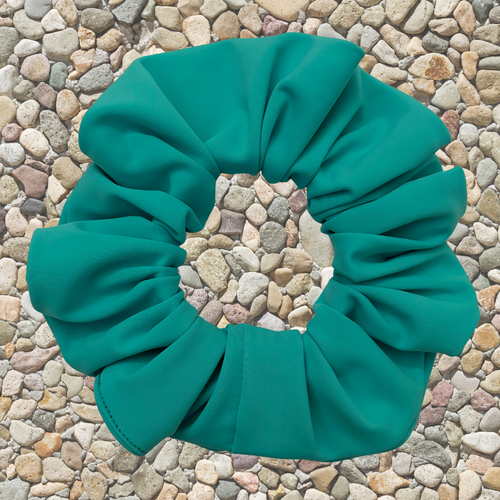 A teal green fabric hair scrunchie laid flat on a background of small multicolored pebbles.