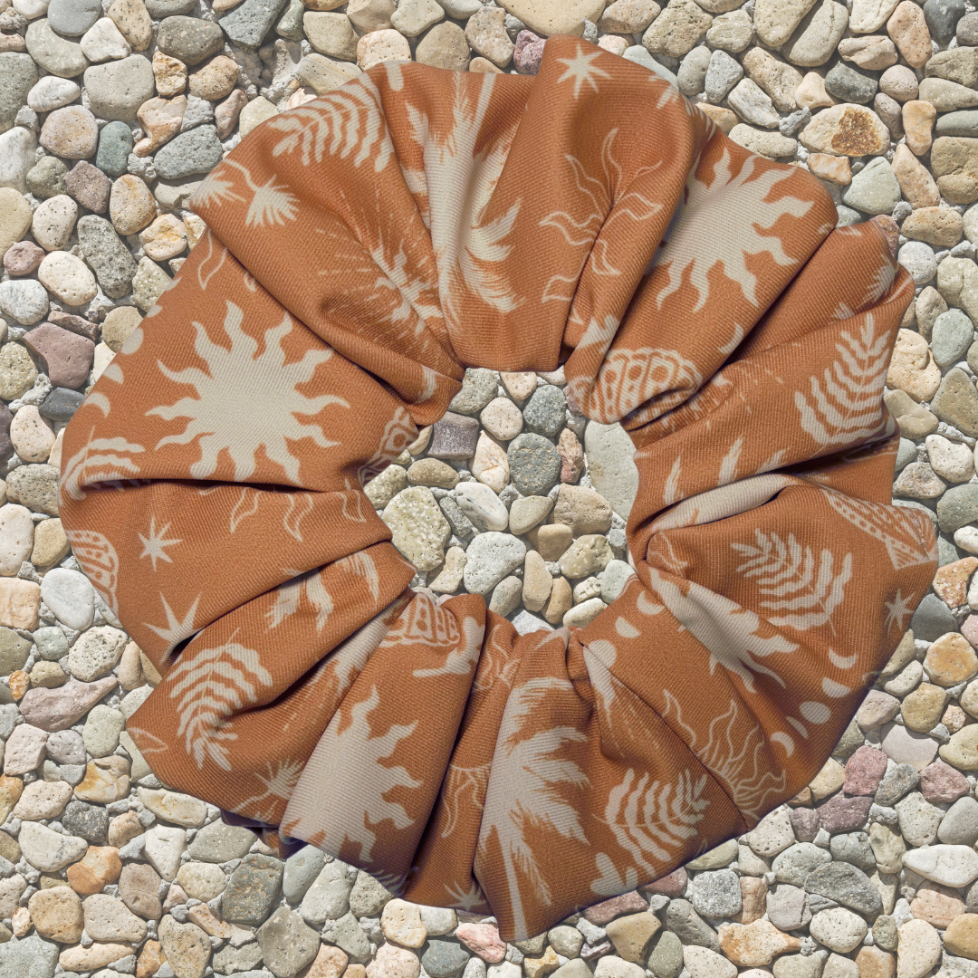 A fabric hair scrunchie with a terracotta orange and cream botanical sunburst pattern, laid flat on a bed of small gray and beige pebbles.