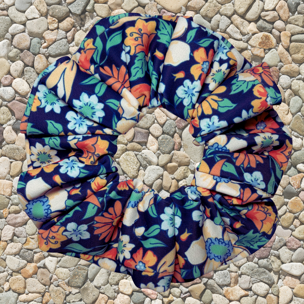A fabric scrunchie with a colorful floral print on a dark navy blue background, placed on a bed of small gray and beige pebbles. The floral pattern features orange, light blue, green, and cream flowers.