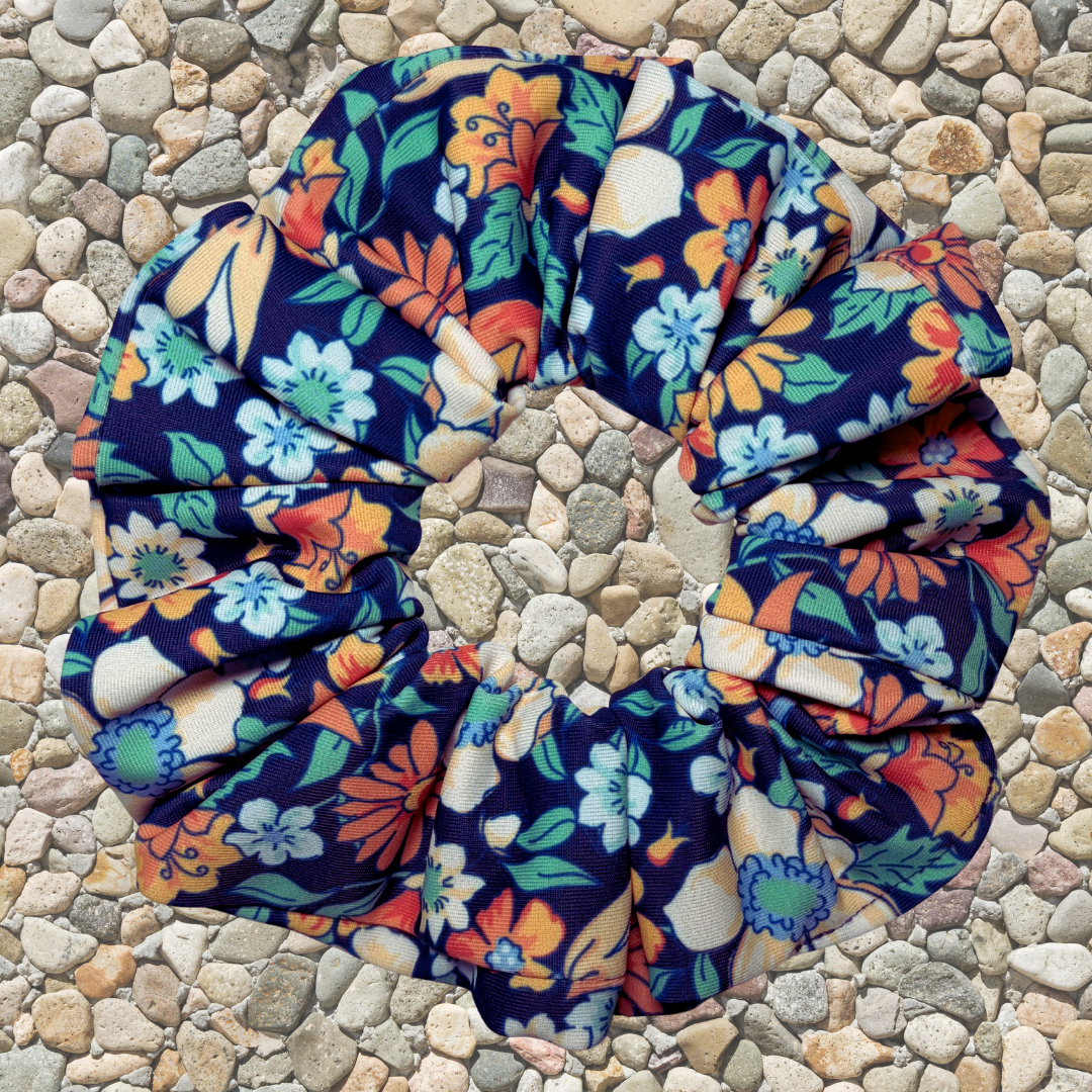 A fabric scrunchie with a colorful floral print on a dark navy blue background, placed on a bed of small gray and beige pebbles. The floral pattern features orange, light blue, green, and cream flowers.
