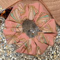 Pink seashell swim scrunchie sitting on hat with sunglasses on pebble surface 