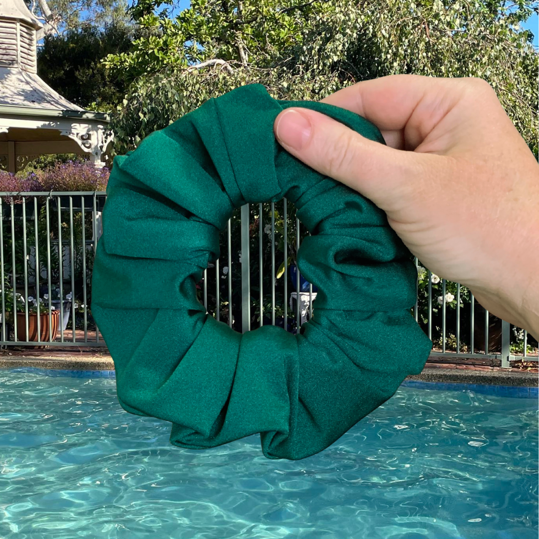 Emerald Green Swim Scrunchie held by a hand with a pool and garden in the background