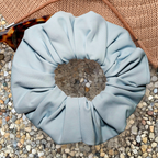 A pale blue fabric scrunchie photographed from above with a brown hat and tortoiseshell sunglasses partially visible.