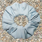 A light blue fabric hair scrunchie laid flat on a bed of small multicolored pebbles.