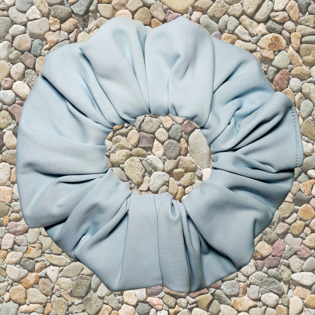 A light blue fabric hair scrunchie laid flat on a bed of small multicolored pebbles.