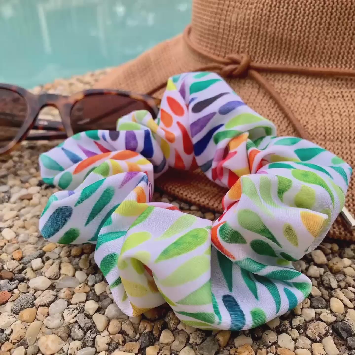 Colourful swim scrunchies on a straw hat with sunglasses on pebbles by a pool.