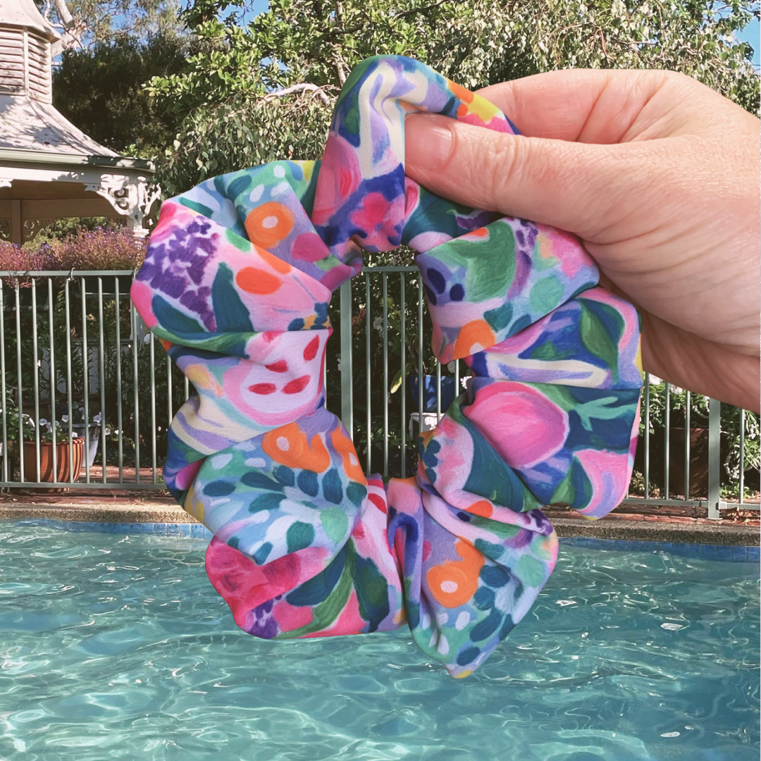 Colourful floral swim scrunchie held above a pool with trees and a building in the background