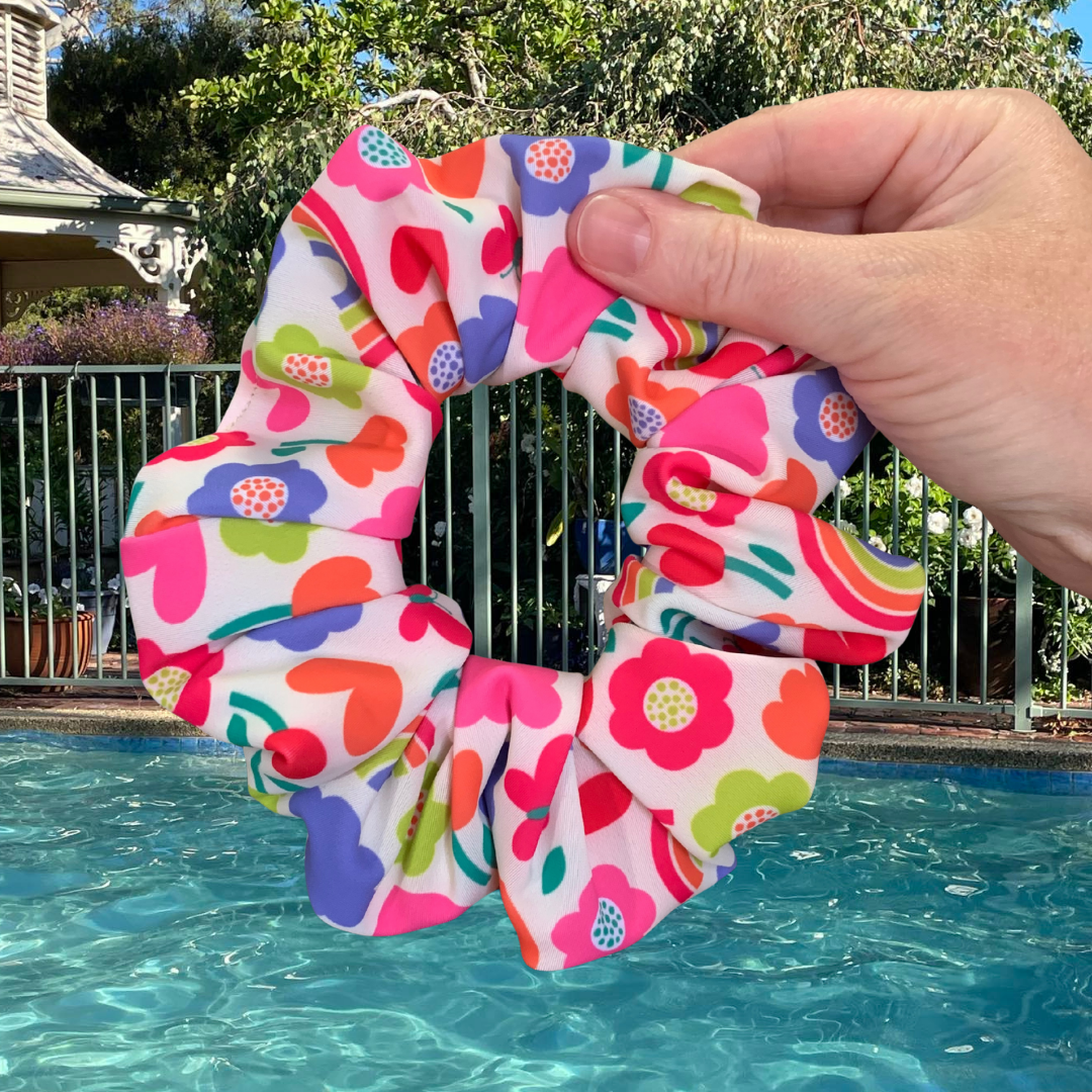 Colourful flower and hearts swim scrunchie held above a pool with a garden background