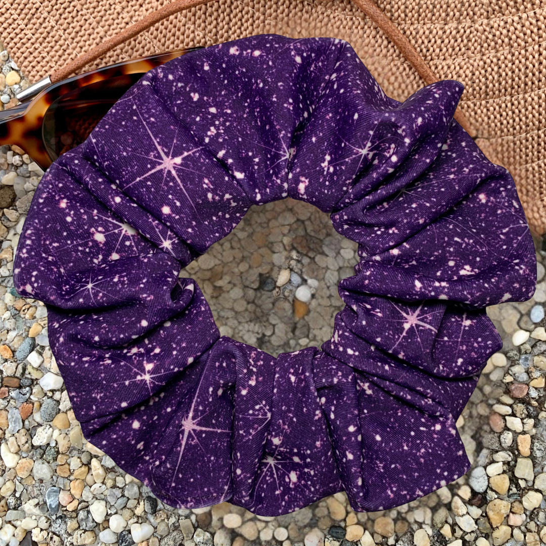Purple printed sparkle swim scrunchie on a textured surface with a hat and sunglasses