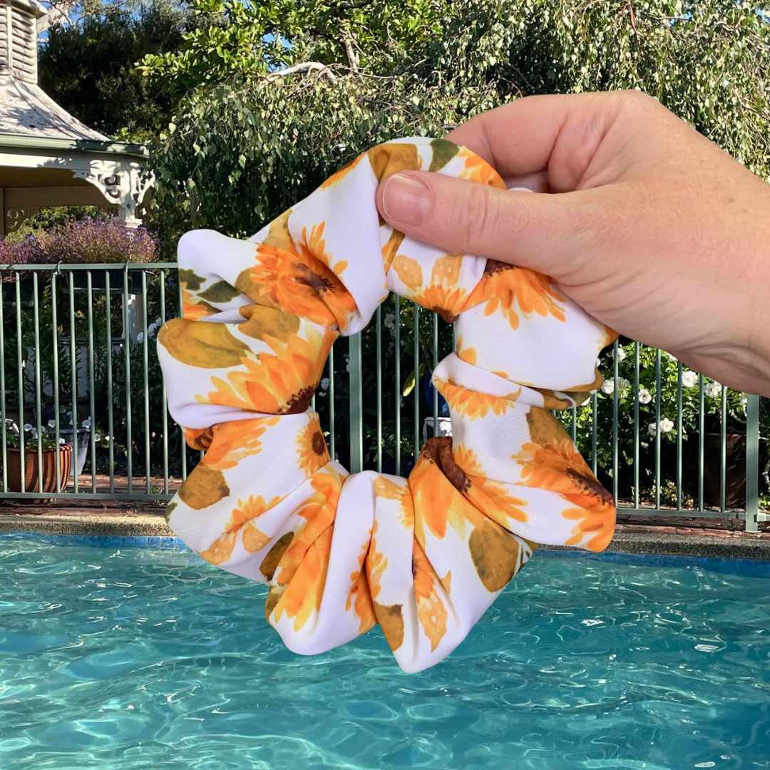 Hand holding a sunflower patterned swim scrunchie with a pool and garden in the background